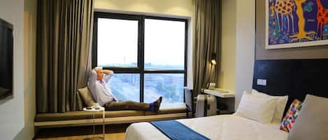 A hotel room with a bed, a window with curtains, a desk, and a painting on the wall.