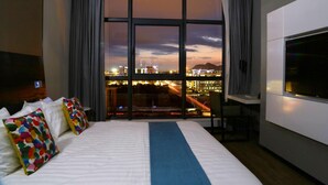 Ein Hotelzimmer mit einem großen Bett, einem Fernseher und Blick auf eine Stadtlandschaft im Abendlicht.