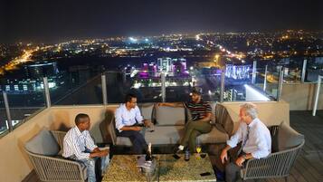 Une terrasse sur le toit où sont assis quatre personnes sur un canapé, une table avec une bouteille et des verres, et une vue sur la ville en arrière-plan.
