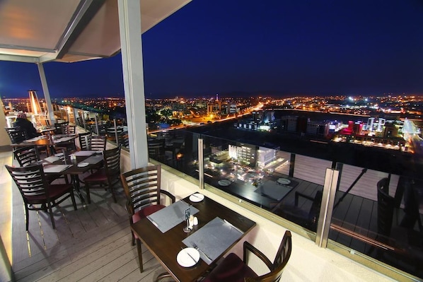 A dining area with tables and chairs, a view of a cityscape at night, and a balcony railing.