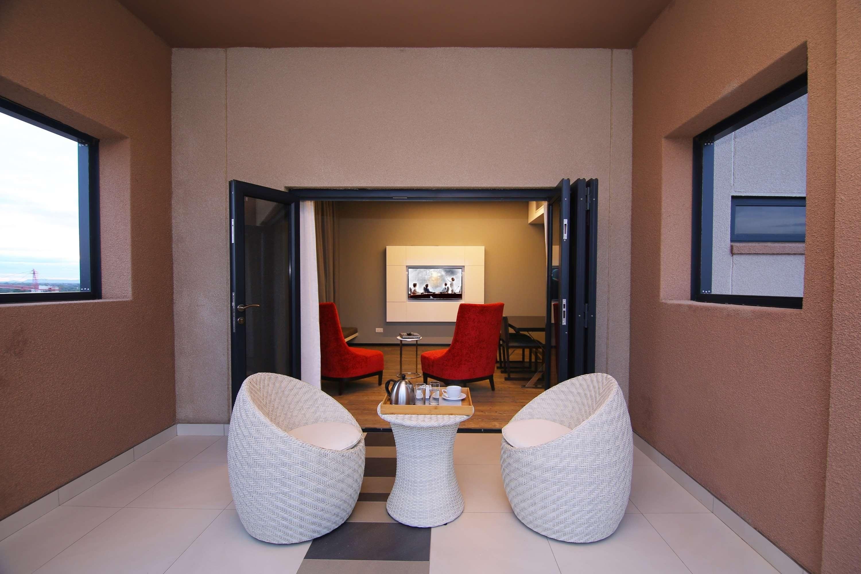 A modern interior with a seating area featuring two white wicker chairs and a small table, a red chair, and a dining table with chairs in the background.