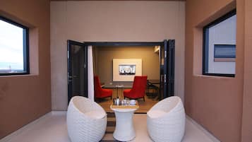 A modern interior with a seating area featuring two white wicker chairs and a small table, a red chair, and a dining table with chairs in the background.