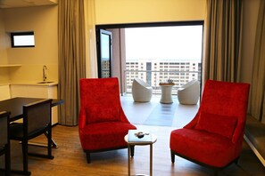 A hotel room with a desk, two red chairs, a small table, a view of the city through a large window, and a white wall-mounted TV. - Room50two (Gaborone)