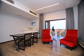 Habitación de hotel moderna con comedor, dos sillones rojos, una mesita y un ventanal con vistas.