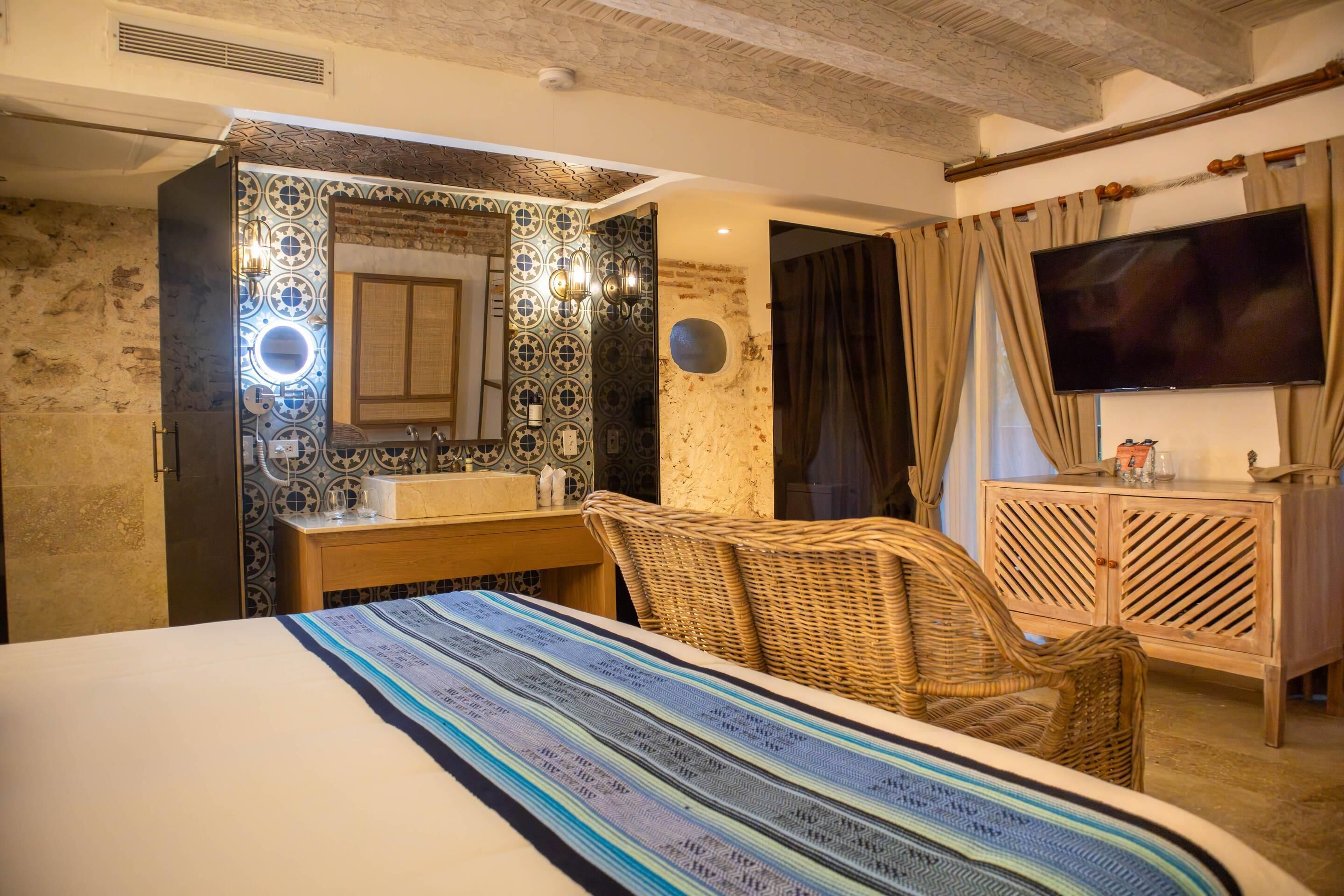 A hotel room with a bed, a wicker chair, a wooden cabinet, a TV, and a bathroom with a tiled wall and a mirror.