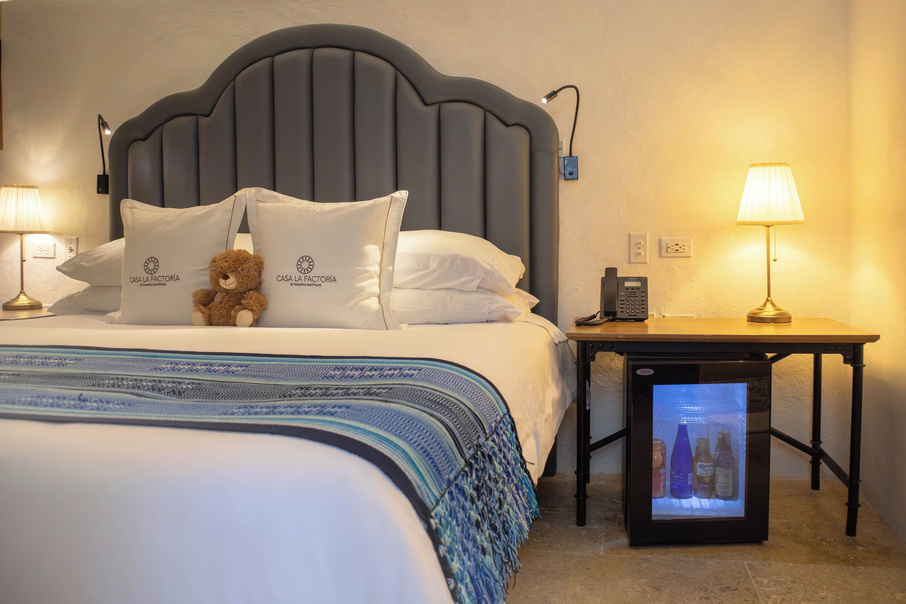 A hotel room with a bed, pillows, a teddy bear, bedside lamps, a nightstand, and a mini-fridge.
