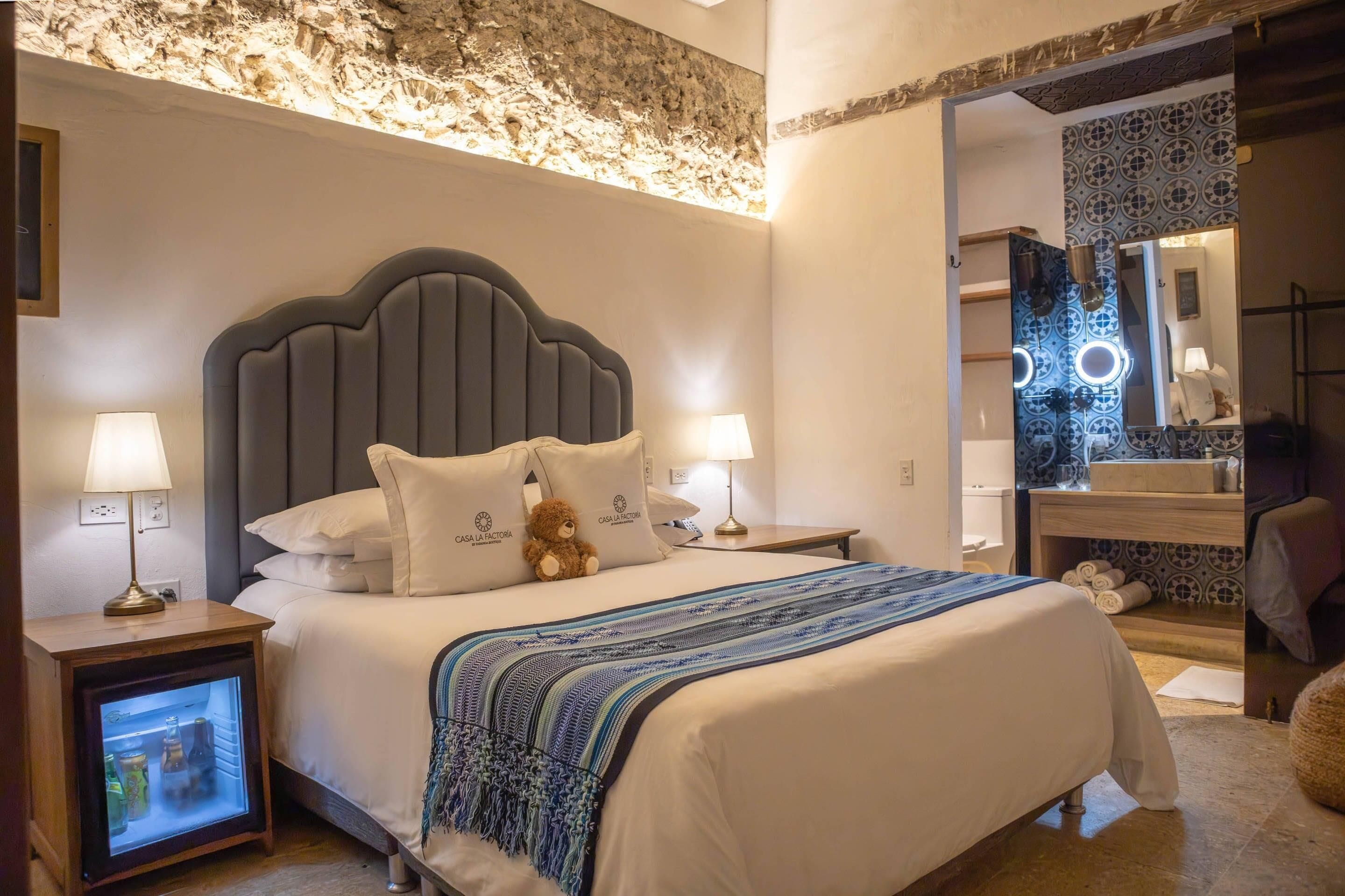 Una habitació d'hotel amb un llit, tauletes de nit, un os de peluix, un mirall i una paret amb un disseny texturat.