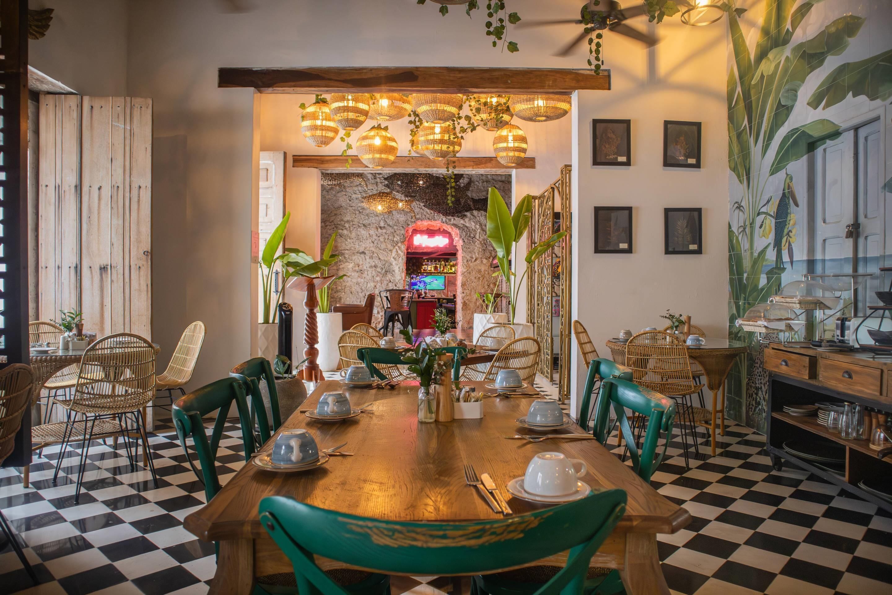 A dining area with a checkered floor, wooden tables, wicker chairs, hanging lights, and a stone wall in the background.