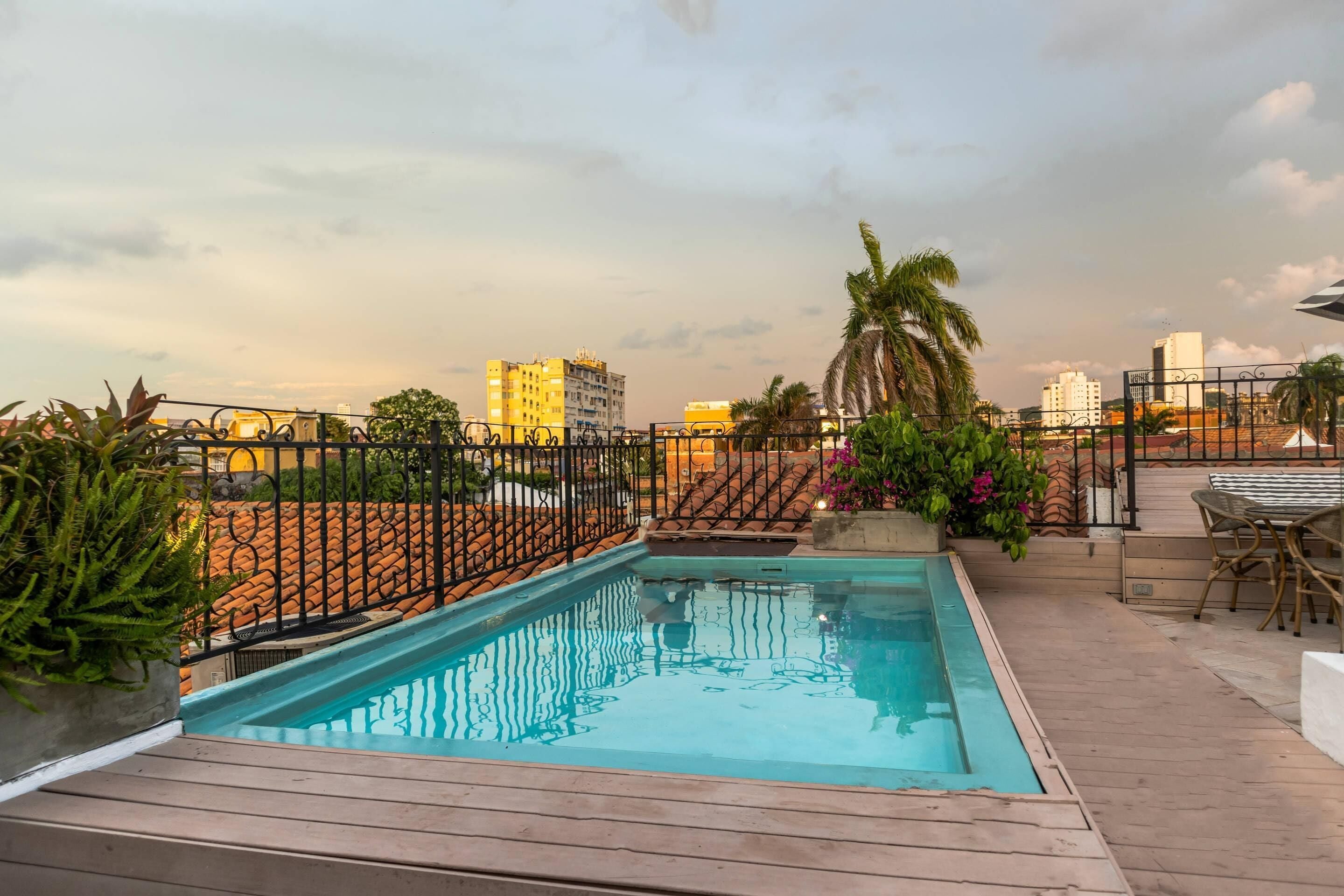 A rooftop pool with a wooden deck, surrounded by a metal fence, with buildings in the background.