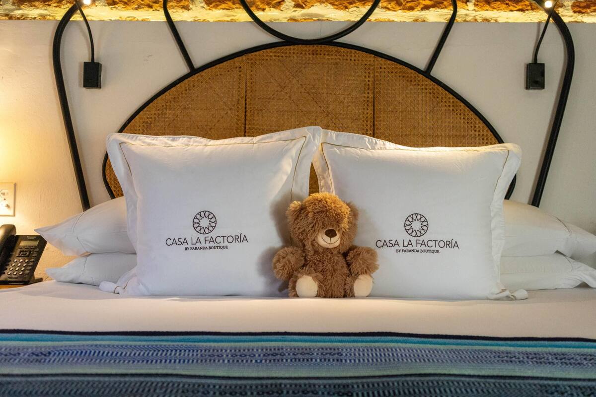 A bed with a brown teddy bear, two pillows with 'CASA LA FACTORIA' text, a black telephone, and a patterned headboard.