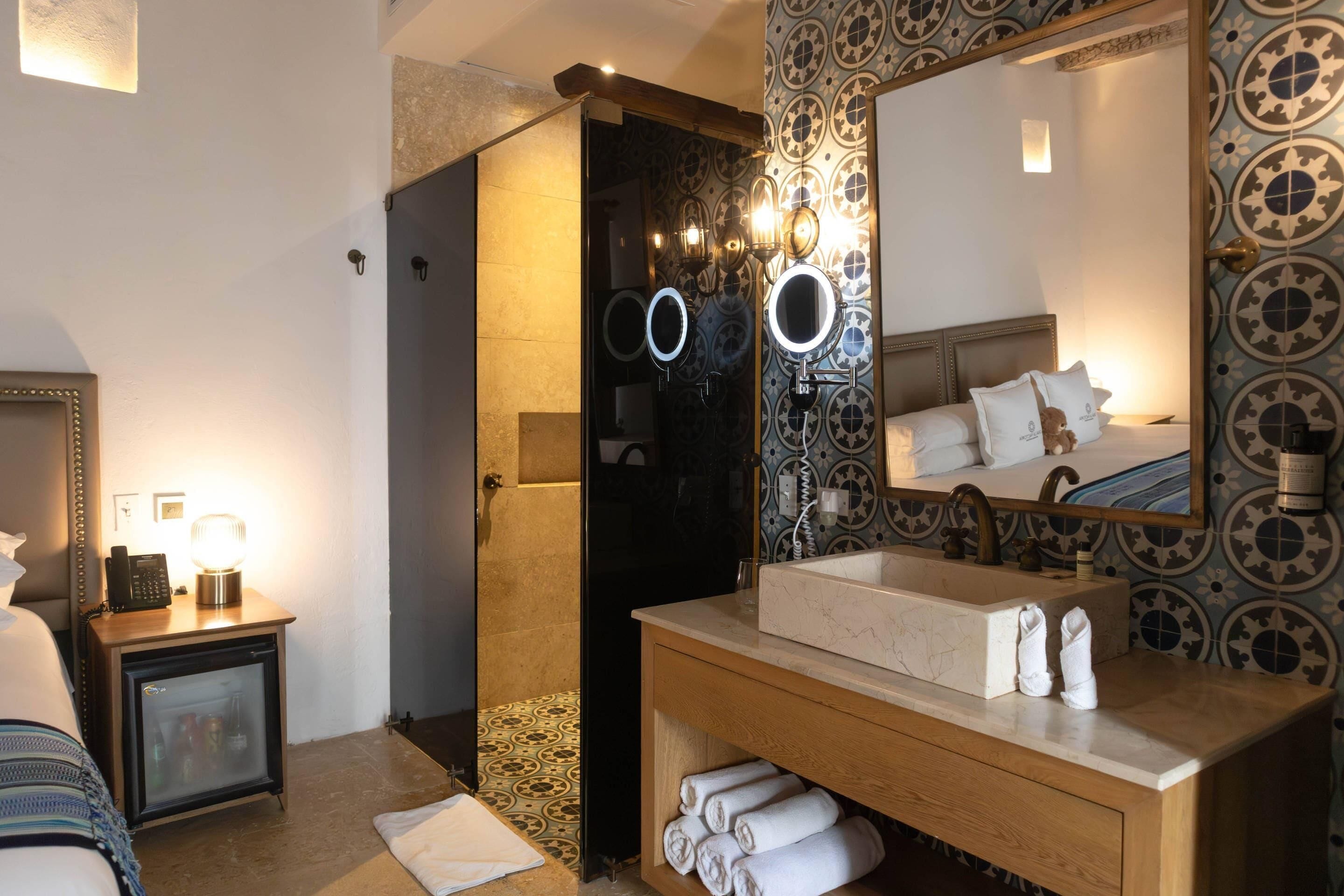 A hotel bathroom with a shower, a sink, a mirror, and a bedside table with a lamp.
