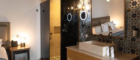 A hotel bathroom with a shower, a sink, a mirror, and a bedside table with a lamp.