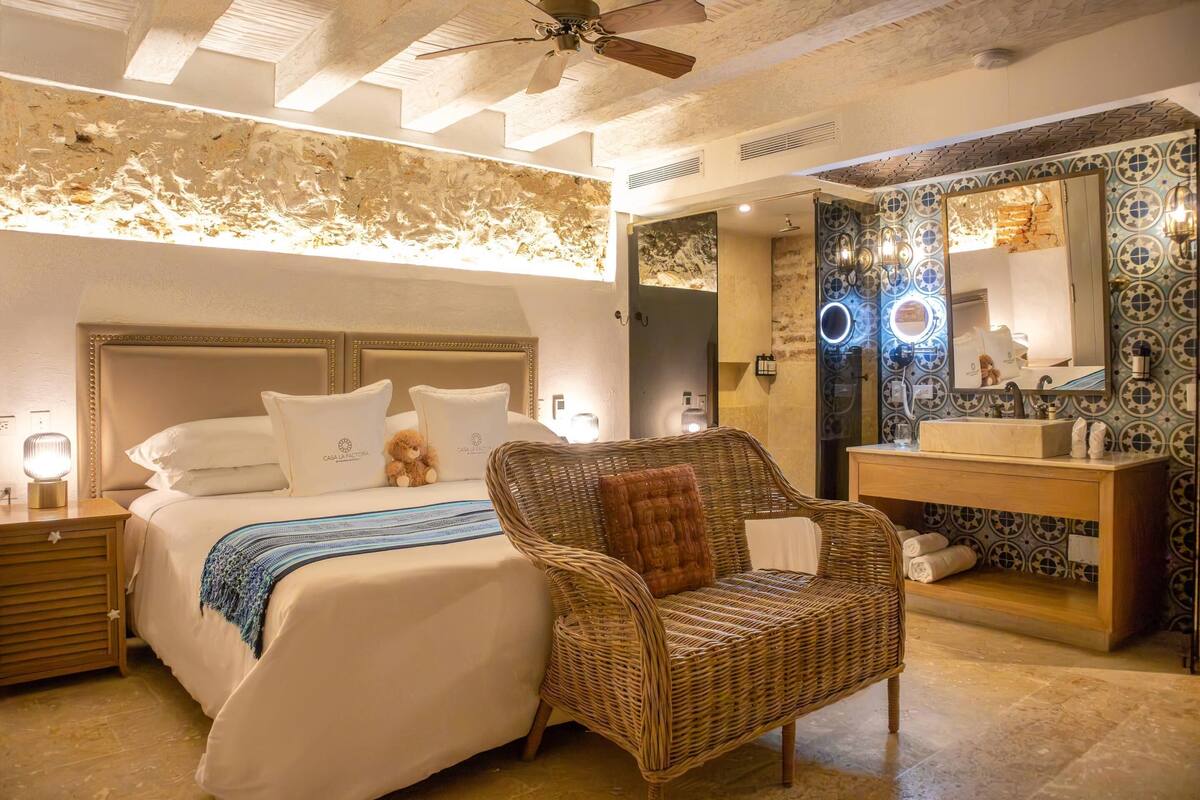 A hotel room with a bed, a wicker chair, a nightstand, a bedside table, a mirror, and a bathroom vanity.