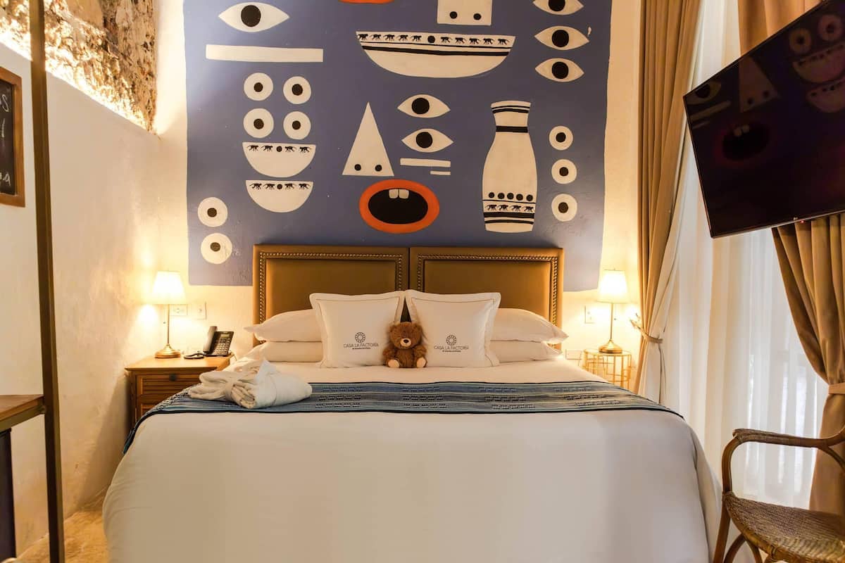A hotel room with a bed, pillows, a teddy bear, a lamp, a nightstand, and a wall with a unique design.