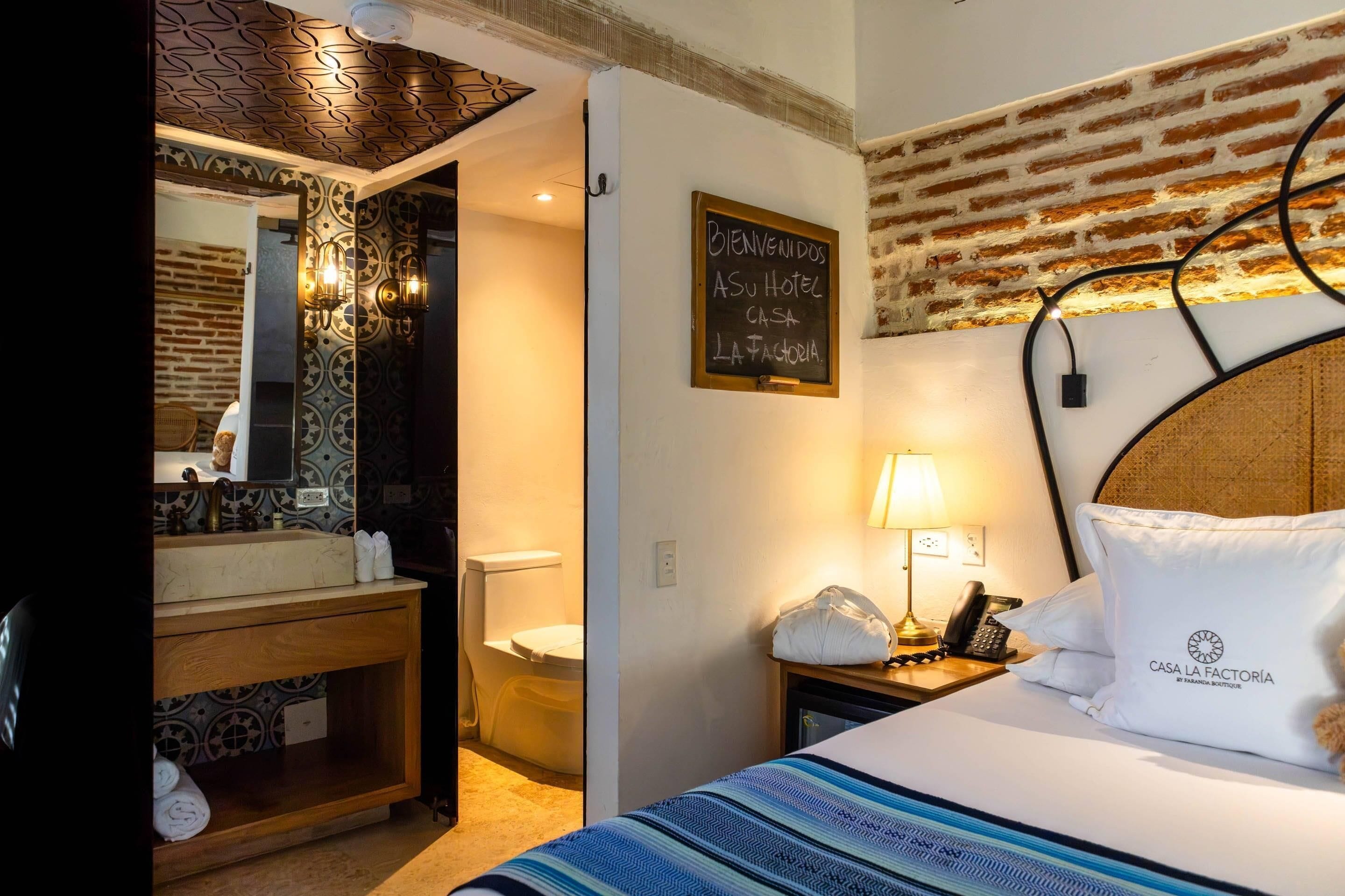 A hotel room with a bed, a nightstand, a lamp, a mirror, a bathroom with a sink and toilet, and a welcome sign.