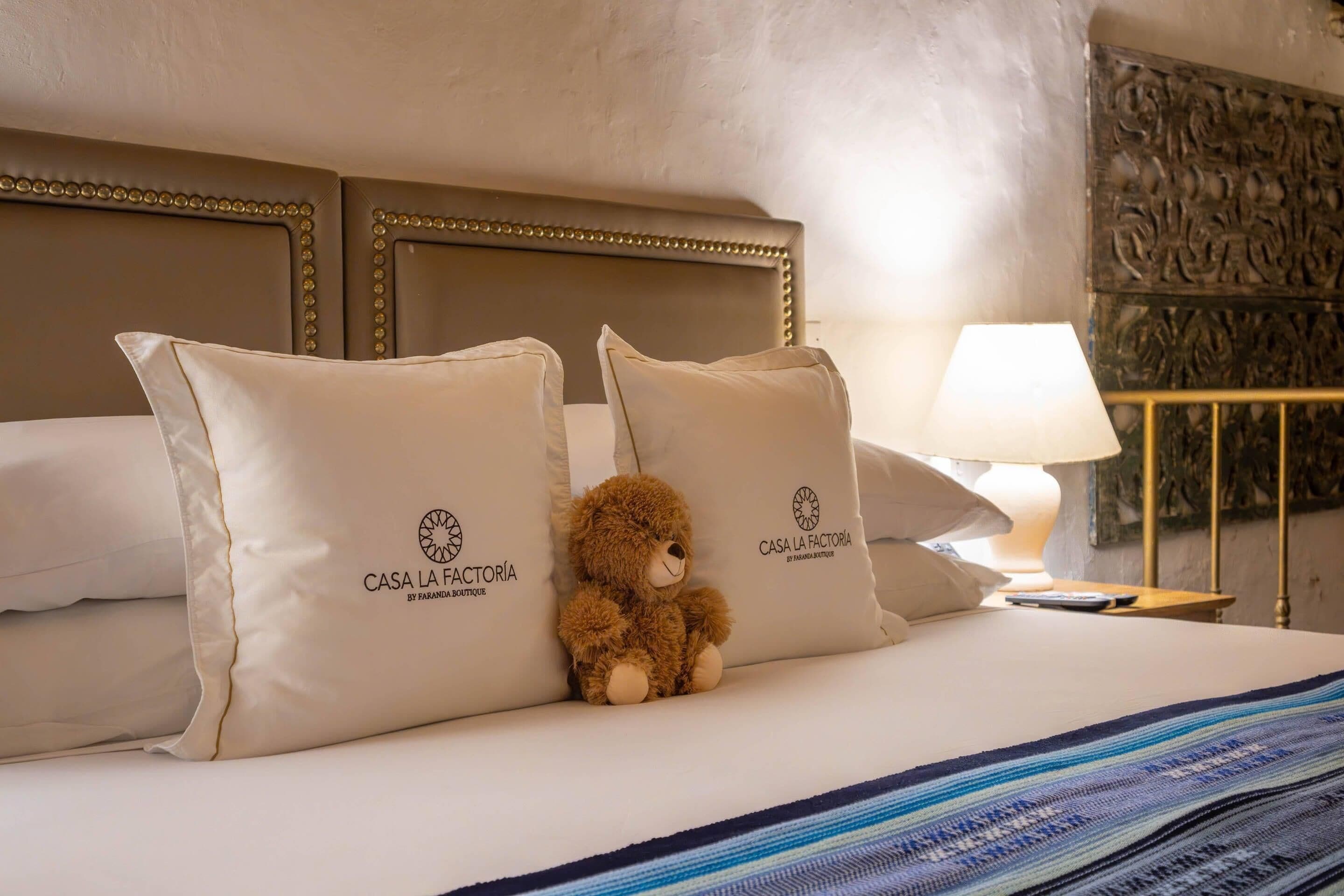 A bed with two pillows labeled 'CASA LA FACTORIA,' a plush teddy bear, a bedside lamp, and a patterned bedspread.