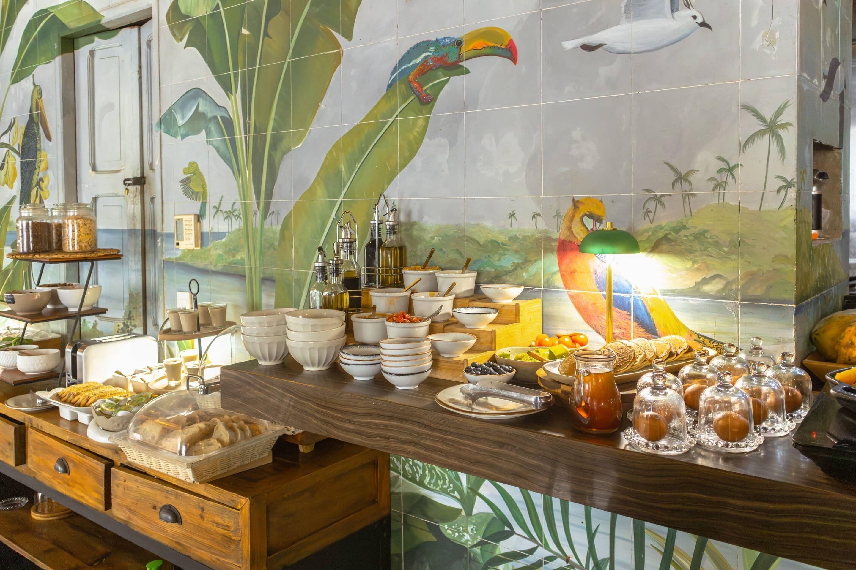A buffet setup with various food items and condiments, a wooden counter, and a mural with tropical birds and foliage.