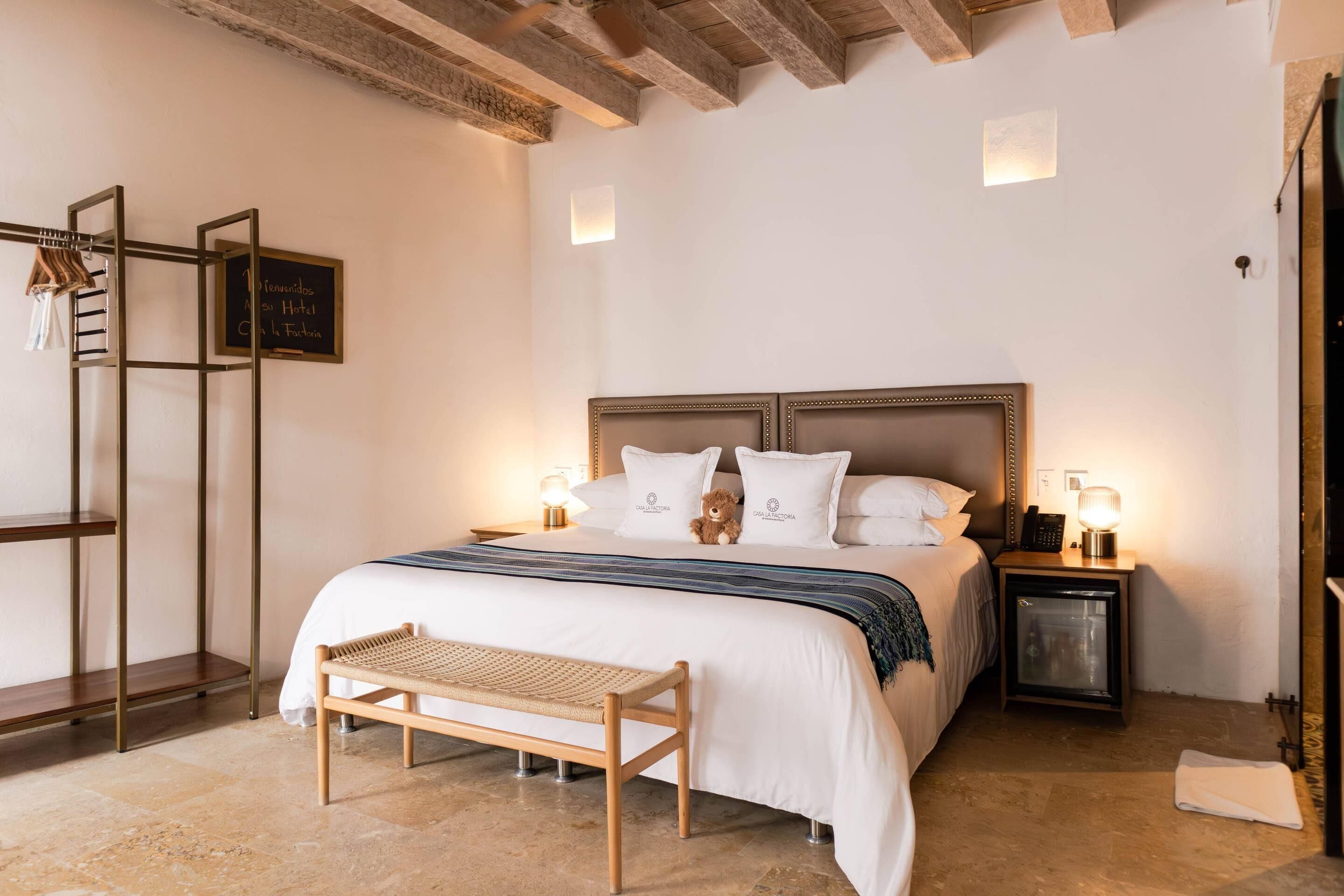 A neatly arranged hotel room with a bed, bedside lamps, a bench, a small table, and a wall-mounted shelf.
