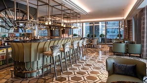Een moderne bar met een hoge plafond, een vloer met een geometrisch patroon, en een uitzicht op de stad door grote ramen.