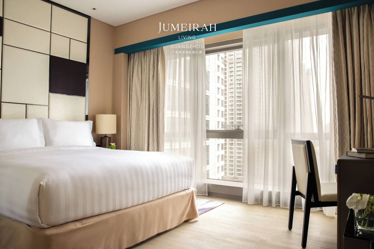 Habitació d'hotel amb un llit gran, un escriptori i vista a alts edificis a través de cortines translúcides.