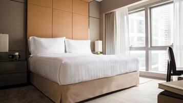 A modern hotel room with a large bed, a nightstand, a lamp, and a view of the city through large windows.