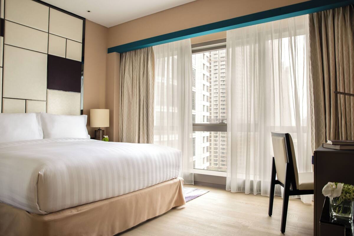 Habitació d'hotel amb un llit gran, un escriptori amb una cadira, una làmpada i vista de campanars a través de cortines primes.