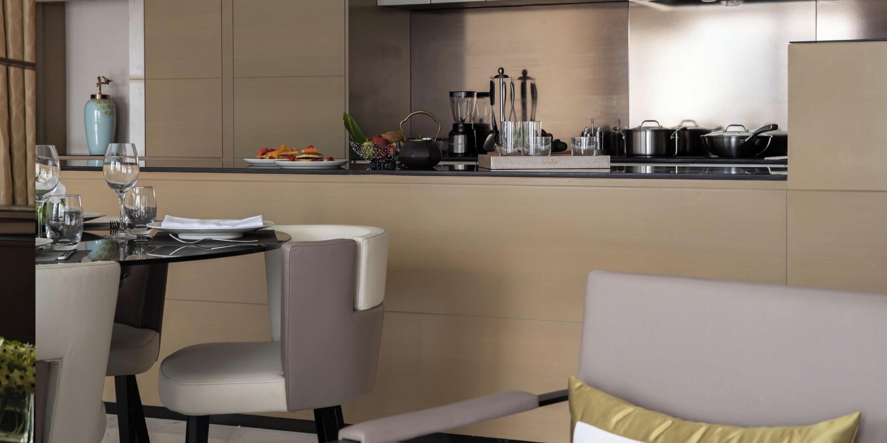 A modern kitchen with a dining area. It includes a table set for two, a chair, and a sofa. Kitchen appliances and utensils are visible on the countertop.