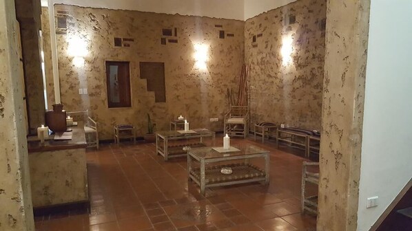 A room with rustic walls, a wooden floor, and a central glass table with candles. There are chairs and a small table with a vase.