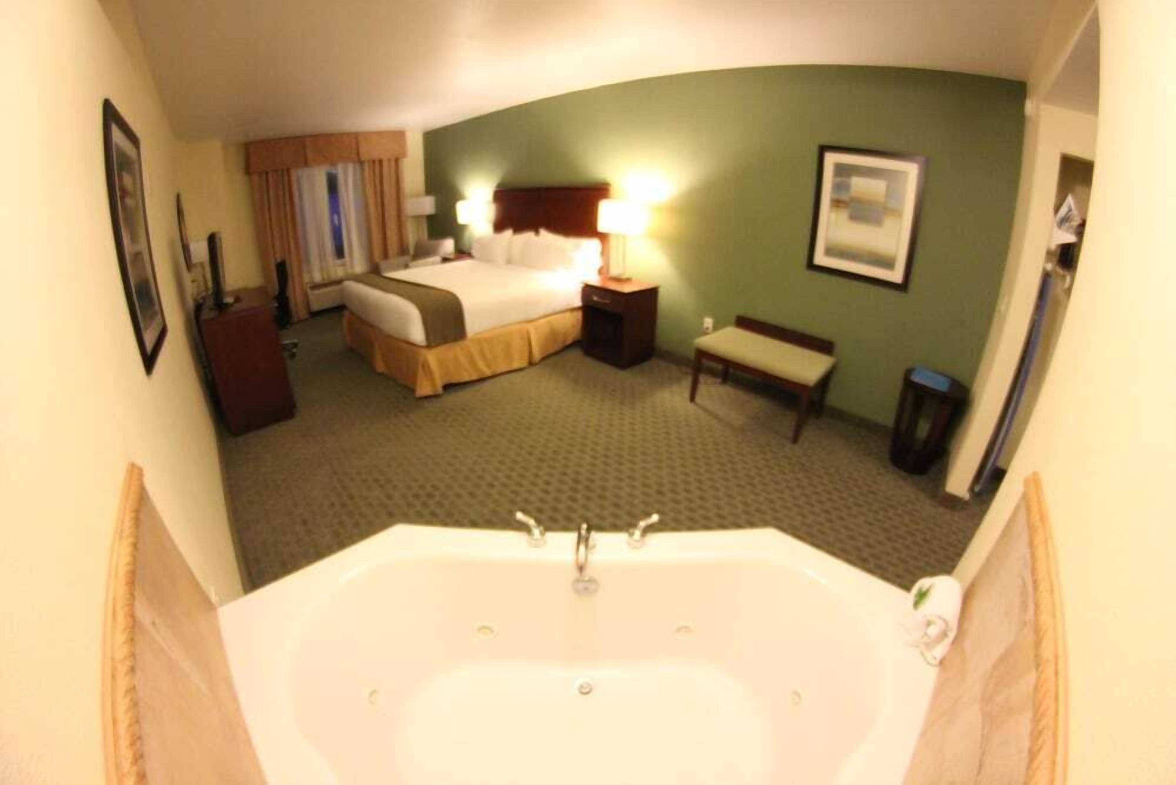 A hotel room with a large bathtub, a bed with a headboard, a bedside table with a lamp, a chair, and a picture on the wall.