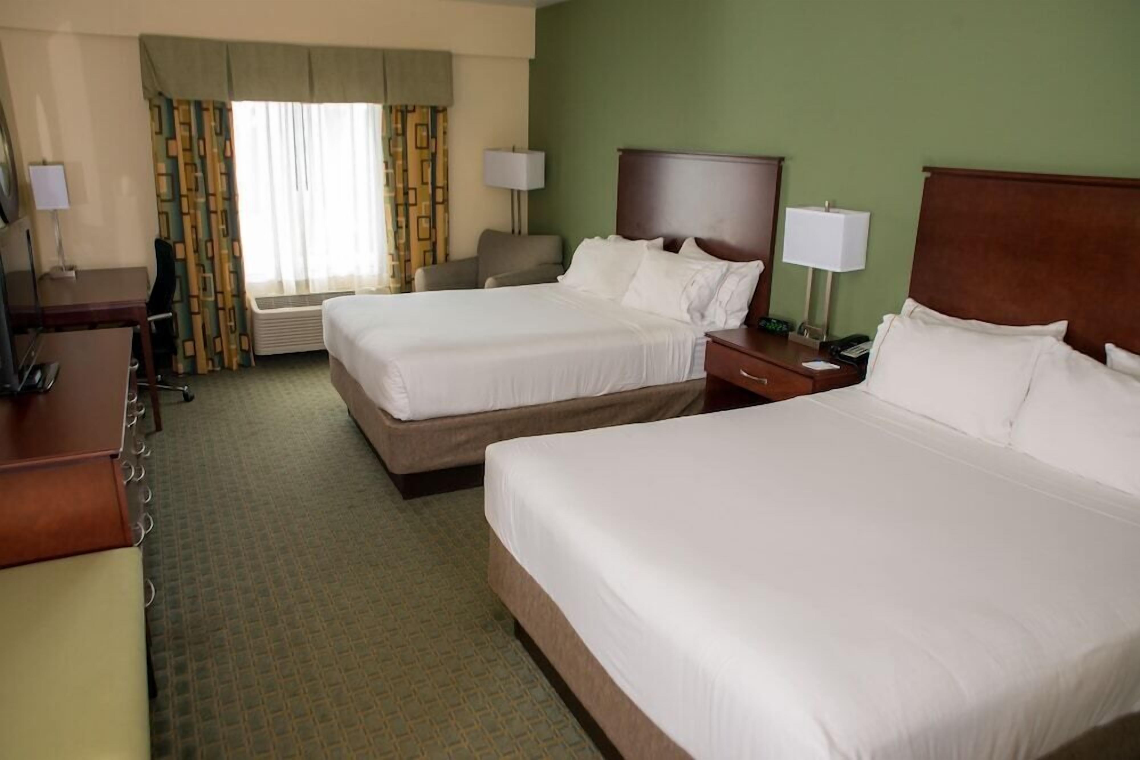 A hotel room with two beds, a desk, a chair, a lamp, and a window with curtains.
