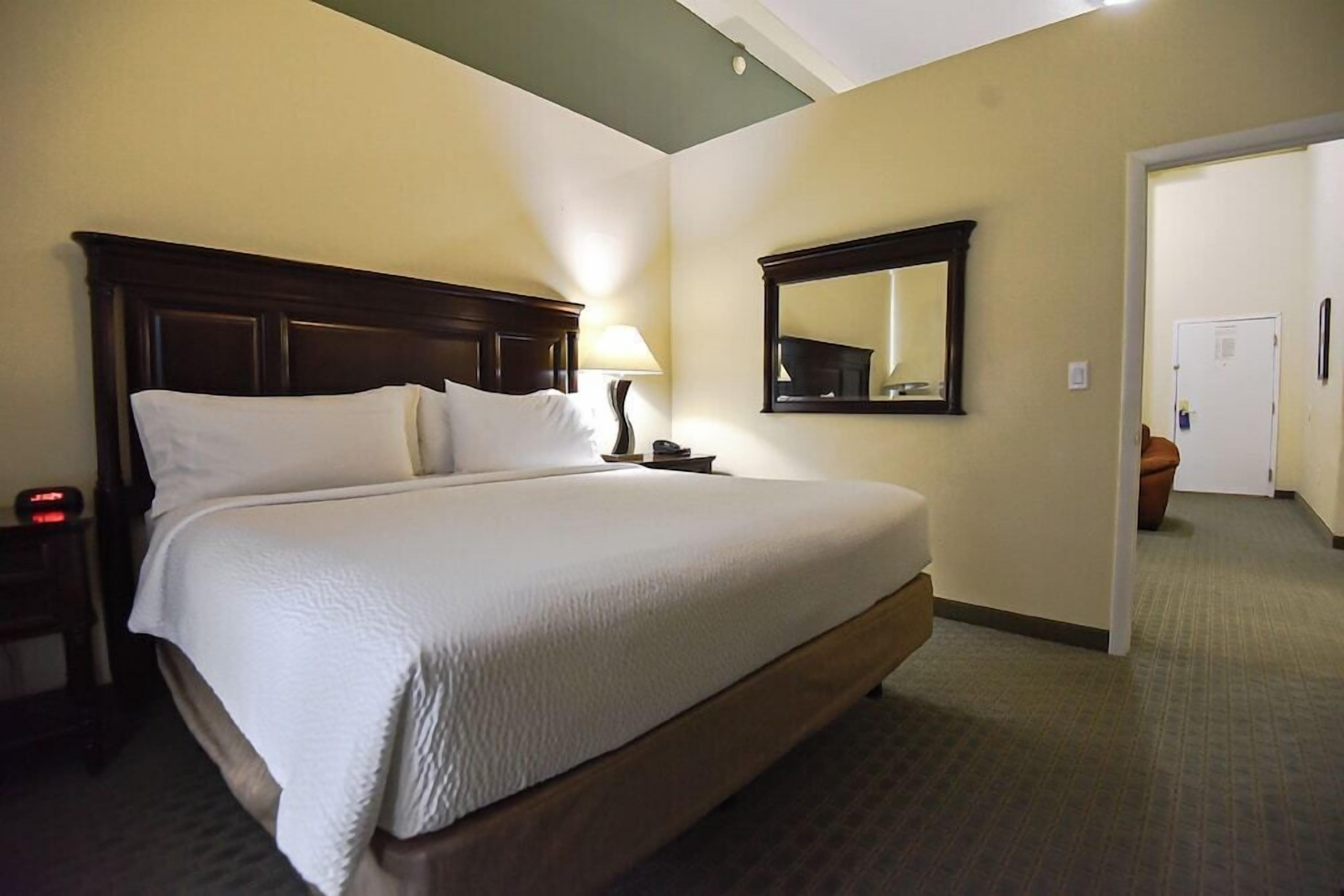 A hotel room with a large bed, a nightstand, a lamp, a mirror, and a door leading to another room.