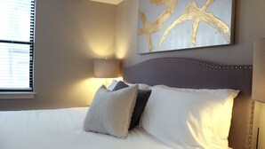 Una cama bien tendida con ropa de cama blanca y gris, un cabecero con tachuelas decorativas y una obra de arte colgada en la pared.