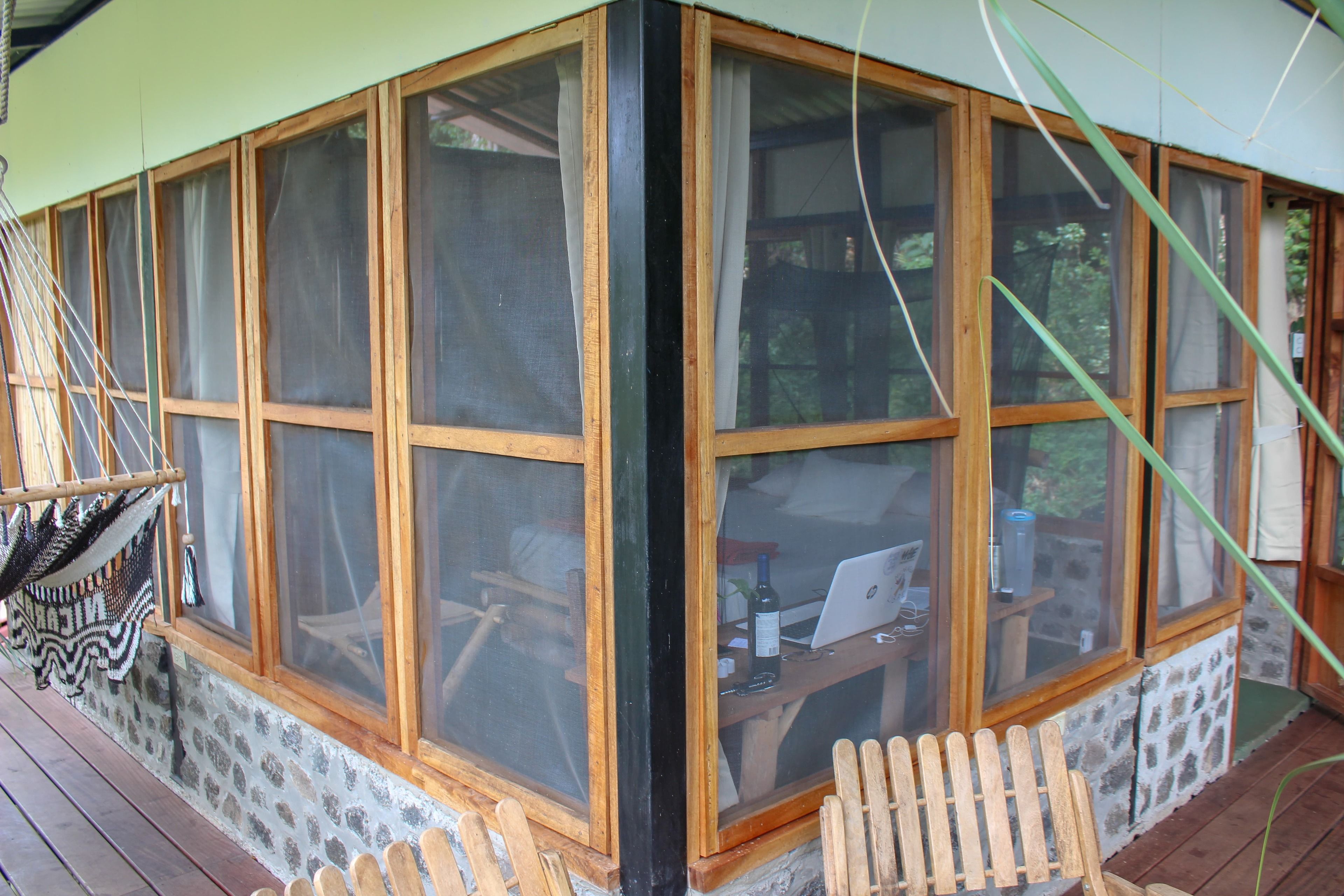A corner room with a large window, a wooden desk with a laptop, and a hammock.