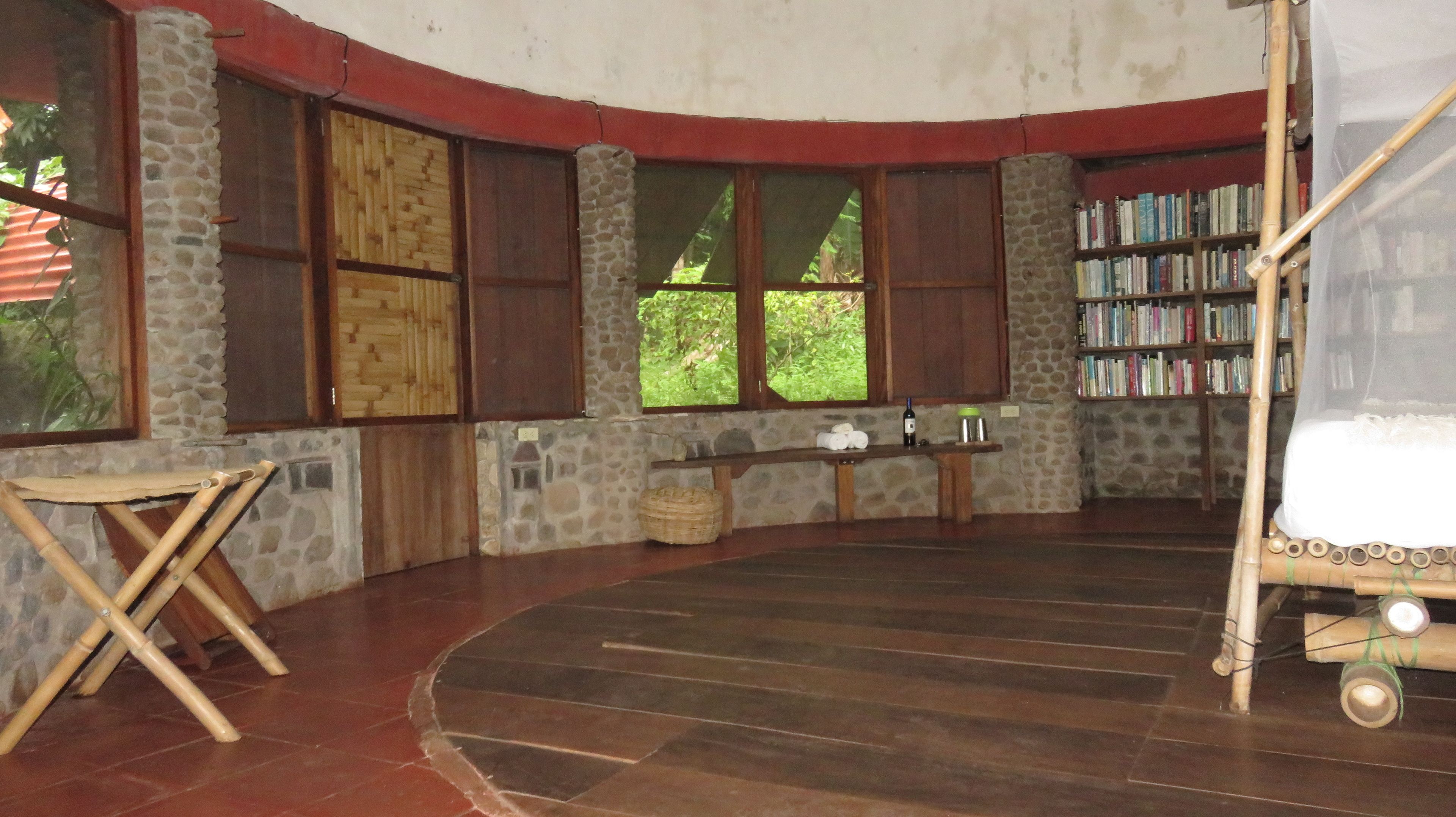 Quarto com parede de pedra curvada, um banco de madeira, uma pequena mesa com uma garrafa de vinho e uma estante de livros.