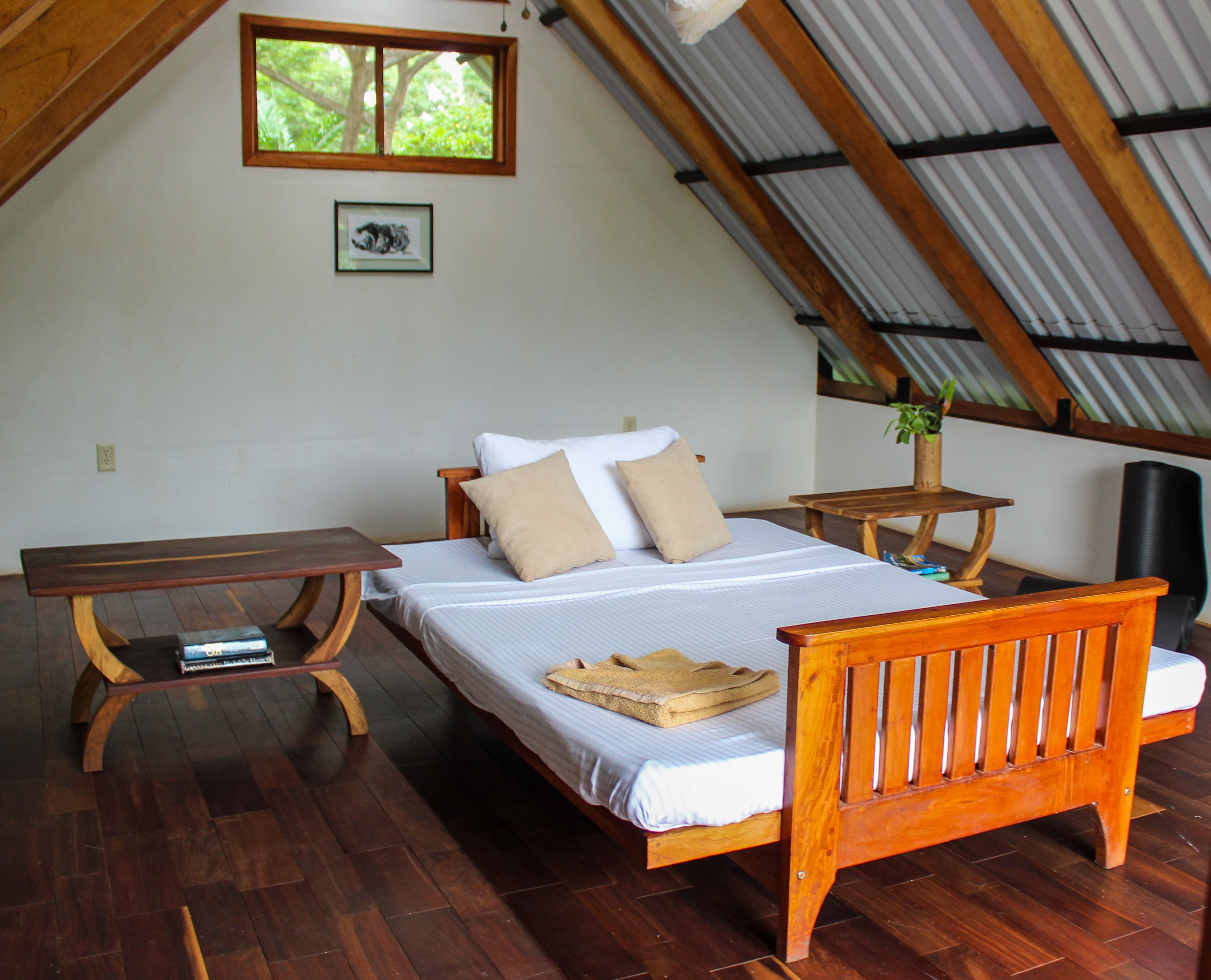 Un dormitorio con una cama y un banco de madera, una mesita y una ventana con vistas a la vegetación.