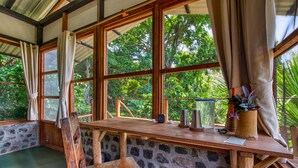Una mesa de madera rústica con un muro de piedra de fondo, amplias ventanas con cortinas y vistas a un follaje exuberante.