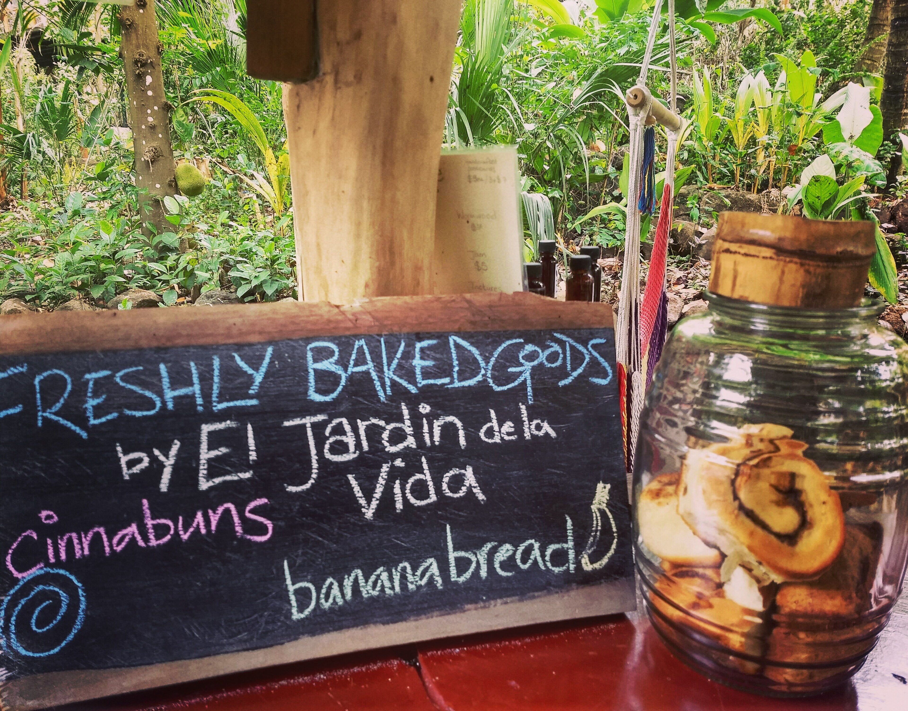 A chalkboard sign listing freshly baked goods, including cinnabuns and banana breads, in a lush, green outdoor setting.