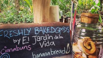 A chalkboard sign listing freshly baked goods, including cinnabuns and banana breads, in a lush, green outdoor setting.