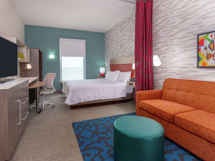 Una habitación de hotel con una cama, un escritorio con una computadora, una silla, un sofá naranja y un pequeño reposapiés.
