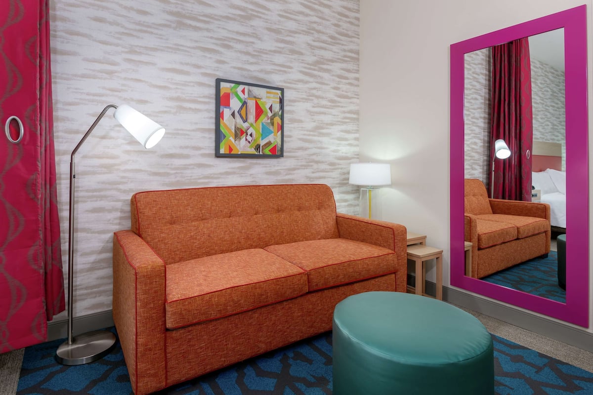 Une chambre d’hôtel avec un canapé, une petite table, une lampe, un miroir et un tableau abstrait aux couleurs vives accroché au mur.