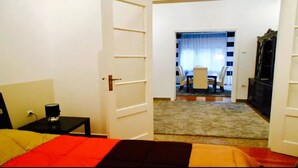 A hotel room with a bed, a bedside table, a lamp, a TV, and a view of a dining area with a table and chairs. - Acropolis detached house (Athens)