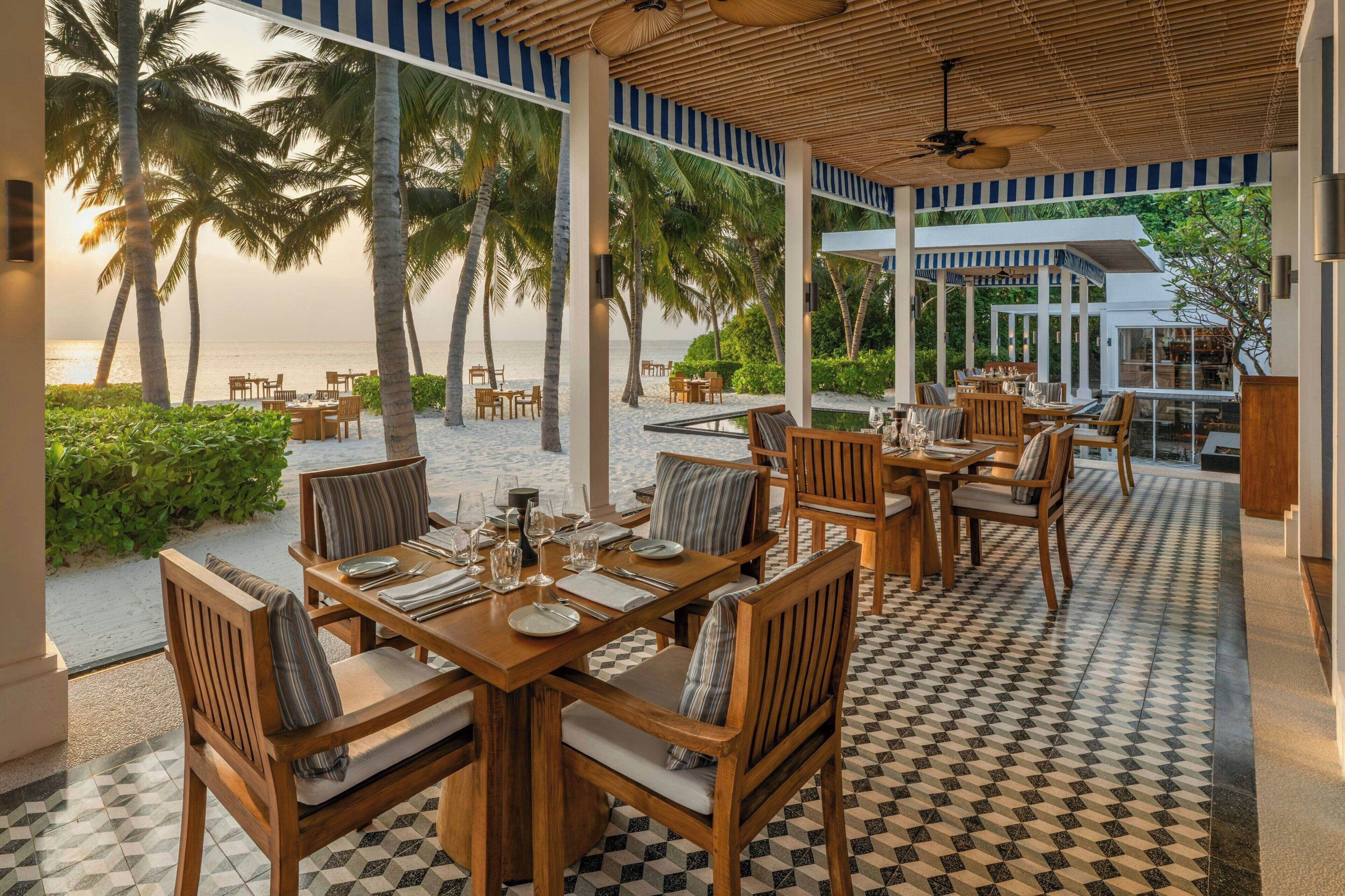 Un restaurante con terraza al aire libre que da a una playa, con mesas y sillas de madera, suelo a cuadros y vistas al océano y palmeras.