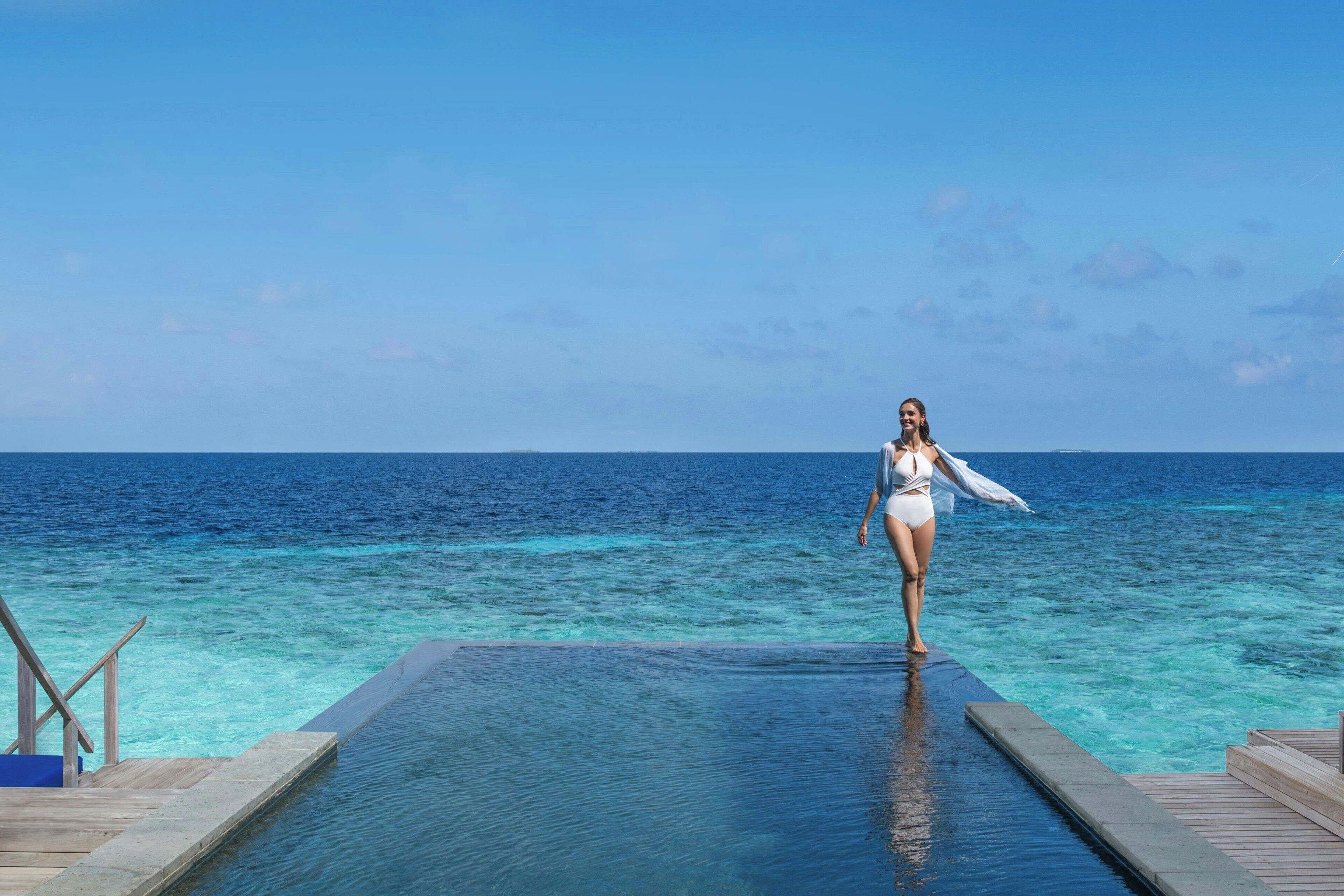 Una mujer caminando sobre una terraza de madera sobre una piscina, con vista al océano.