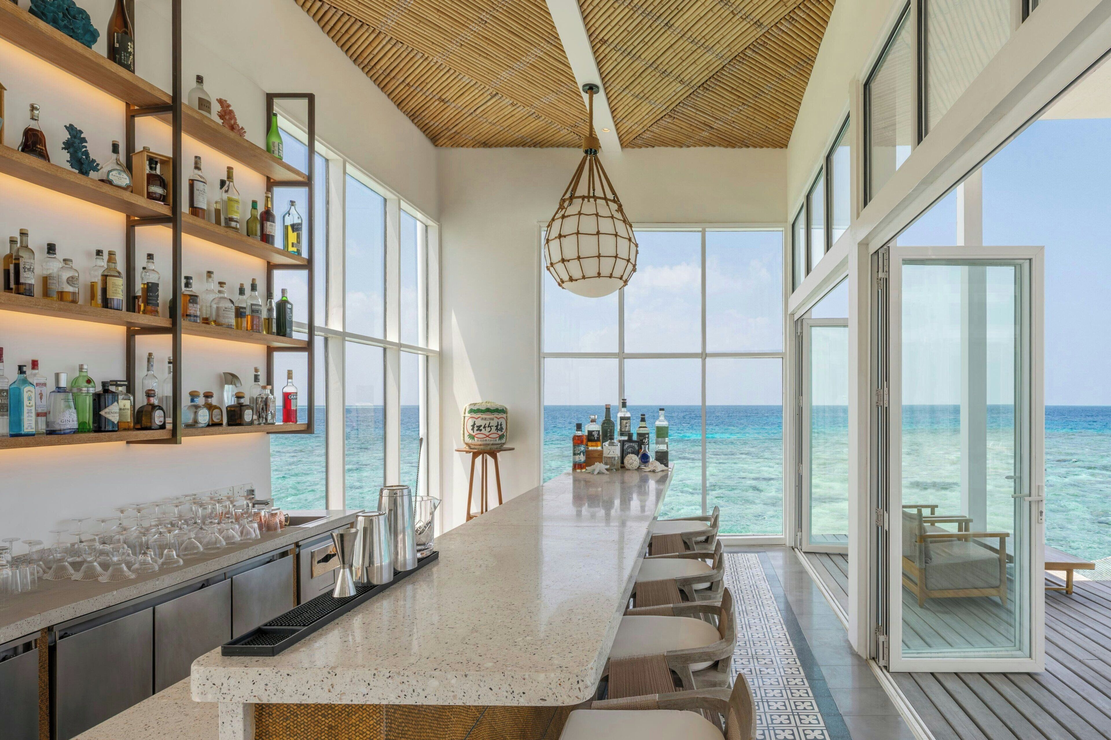Zona de bar con vistas al mar, estantes con botellas y un área de estar también con vistas.