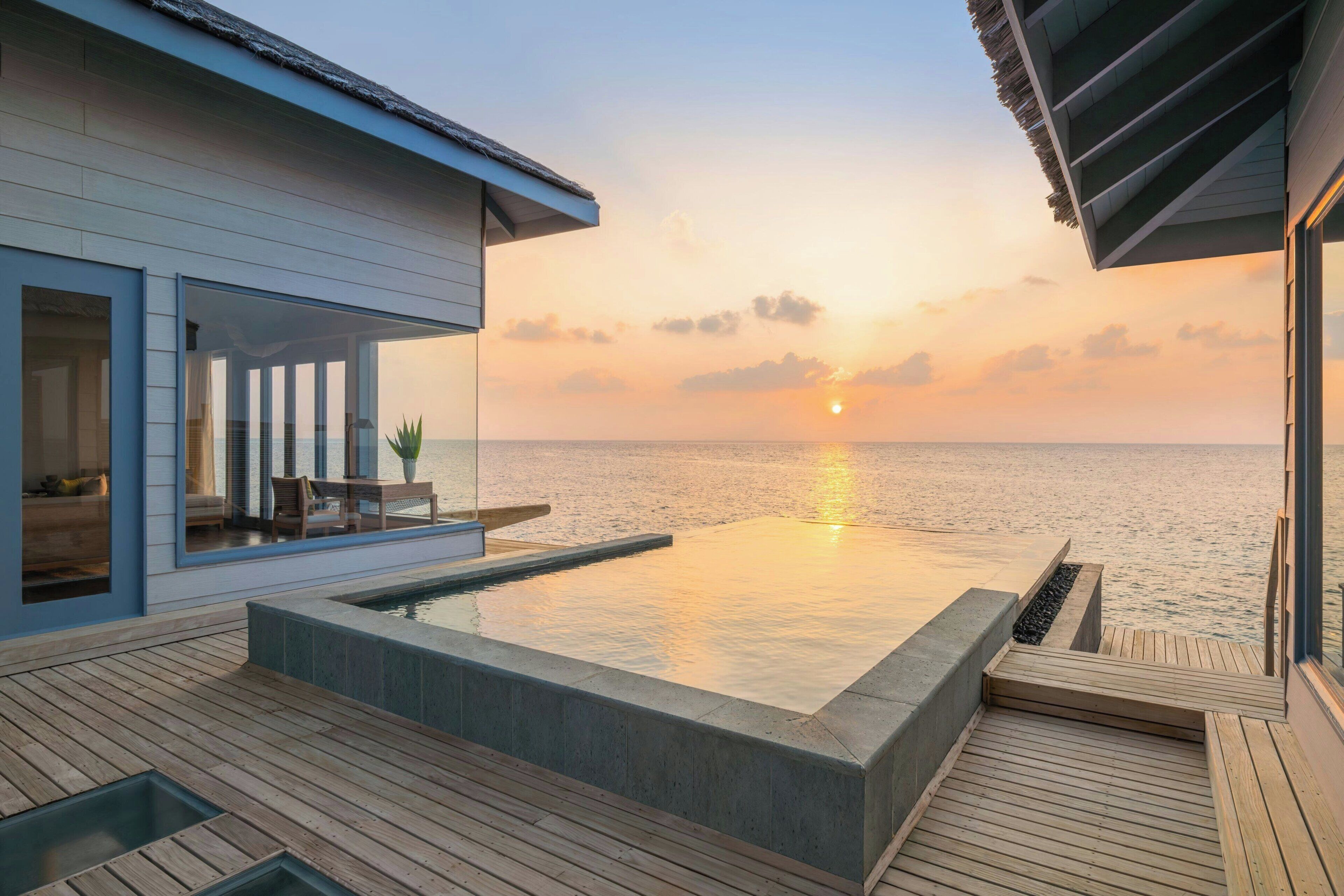 Piscina infinita moderna con vistas al océano al atardecer, terraza de madera y zona acristalada con asientos.