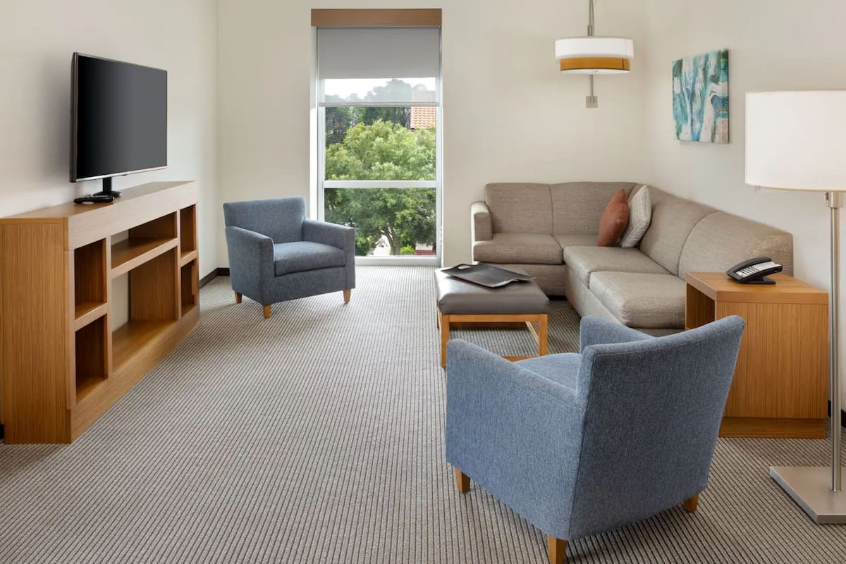 Una habitación de hotel moderna con una televisión de pantalla plana, un sofá, una silla, una mesa de centro, una lámpara, un teléfono y una estantería.