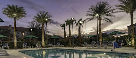 A swimming pool with lounge chairs and umbrellas, surrounded by palm trees and street lamps.