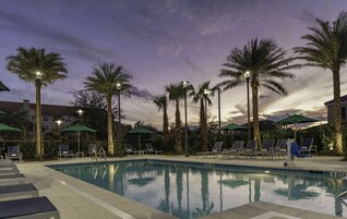 Una piscina con sillones de playa y sombrillas, rodeada de palmeras y farolas.