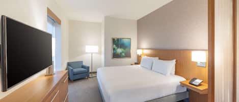 A hotel room with a large flat-screen TV, a bed with white linens, a blue armchair, a painting on the wall, and a bedside table with a lamp and phone.