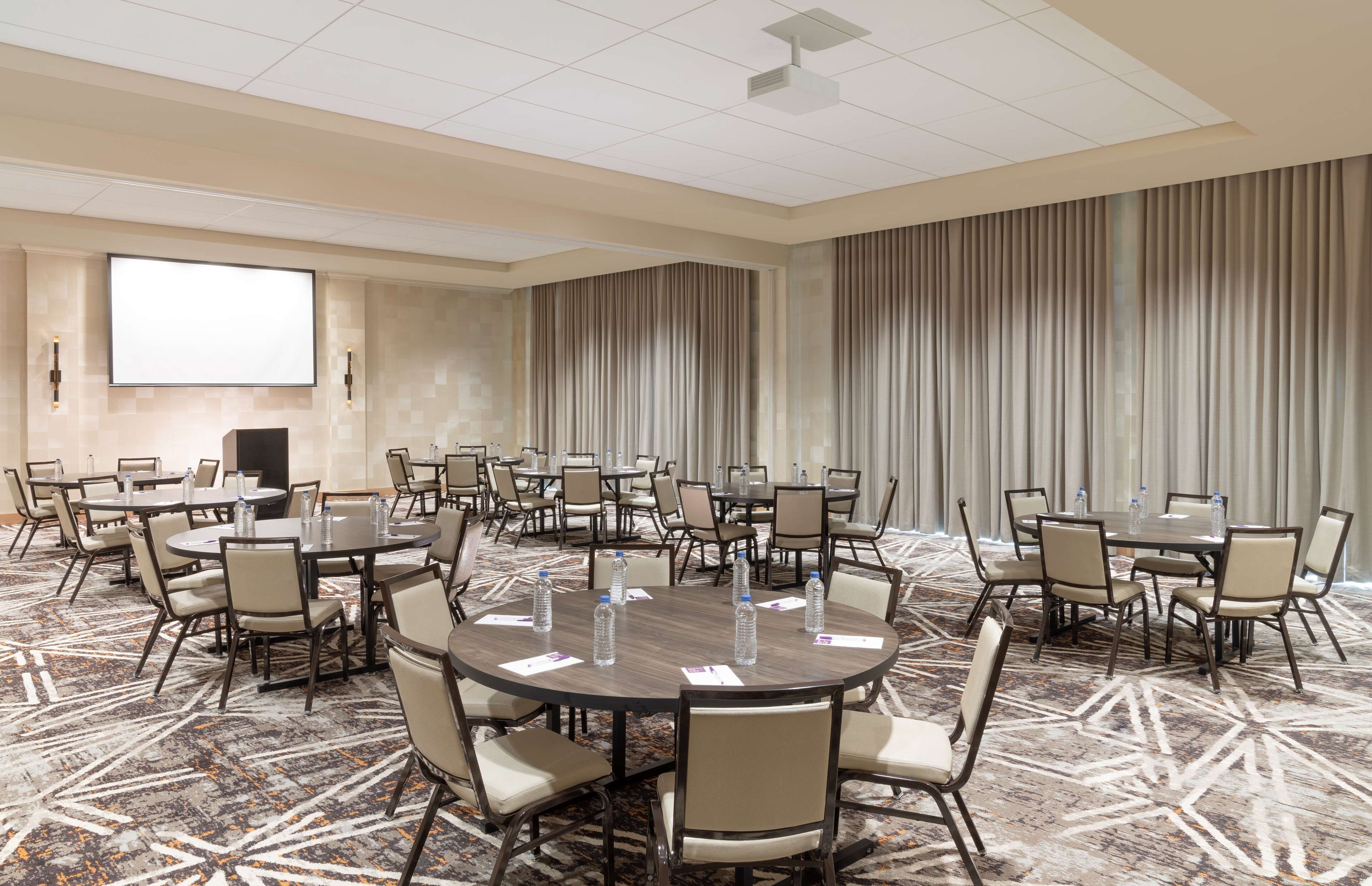 A conference room with multiple tables and chairs, a projector screen, and a large window with curtains.